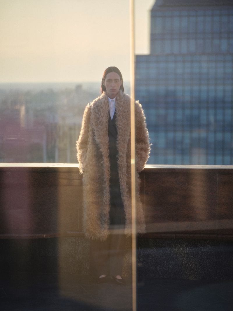 Model in fluffy textured camel coat standing on a rooftop against city skyline at dusk