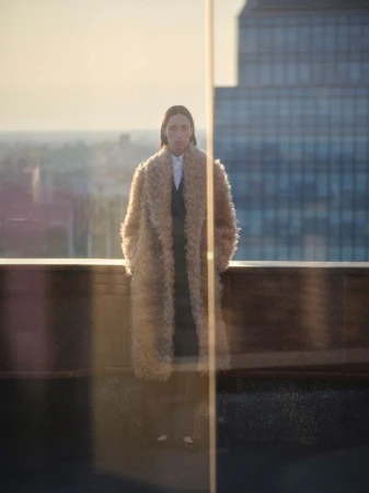 Model in fluffy textured camel coat standing on a rooftop against city skyline at dusk