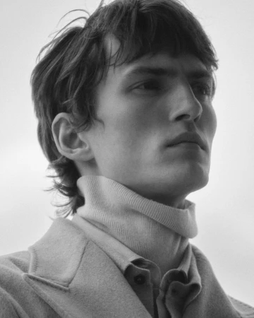 Portrait of a man wearing a high-neck sweater and collared jacket against a pale sky background