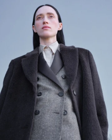 Low-angle portrait of a model with dark hair wearing a heavy grey double-breasted coat against a plain sky blue backdrop