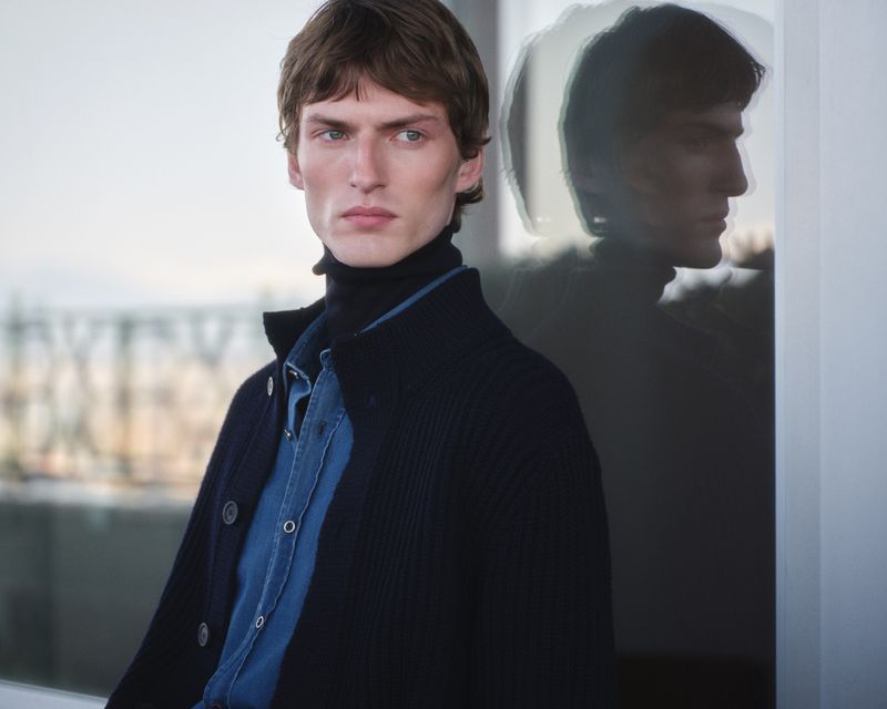 Male model wearing a dark navy cardigan and turtleneck leaning near a window with his reflection visible