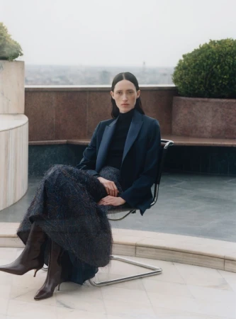 Model Helena Severin seated on a rooftop terrace wearing a dark blazer, turtleneck, and textured skirt