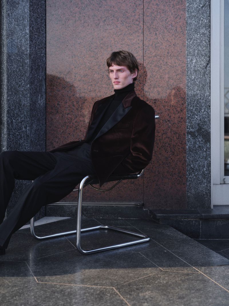 Male model seated in a tubular steel chair wearing a dark velvet blazer and black trousers outdoors