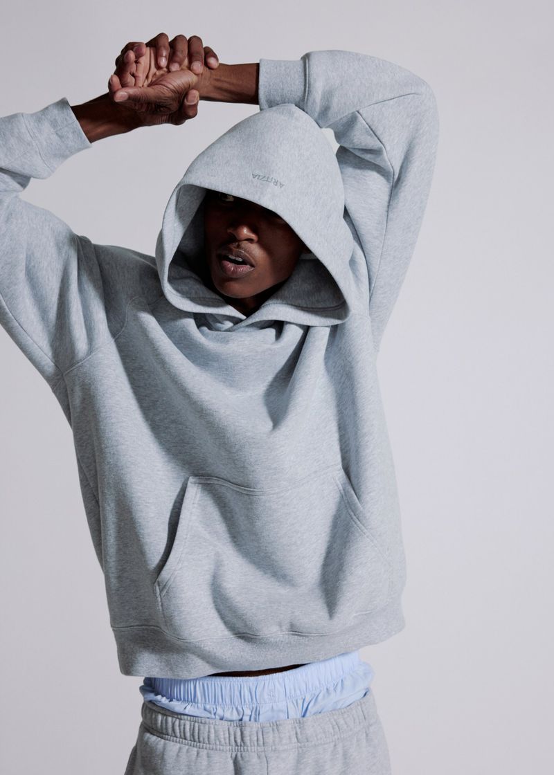 Model wearing a grey hooded sweatshirt stretching arms overhead against a seamless grey backdrop
