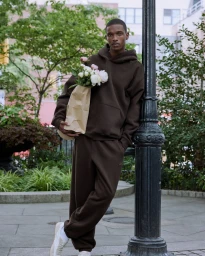Young Black man in a dark brown sweatfleece set leaning against a lamppost and holding a paper bag with flowers