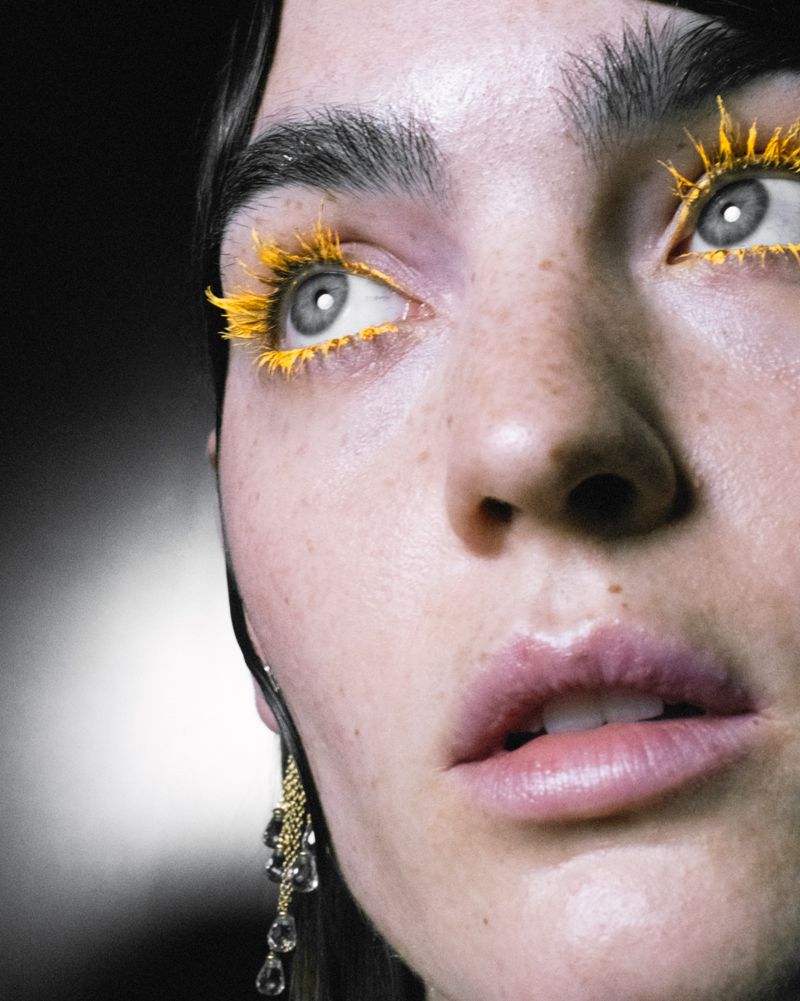 Close-up portrait of a woman with yellow mascara, full lips, freckled skin, and dangling earrings against a dark studio background.