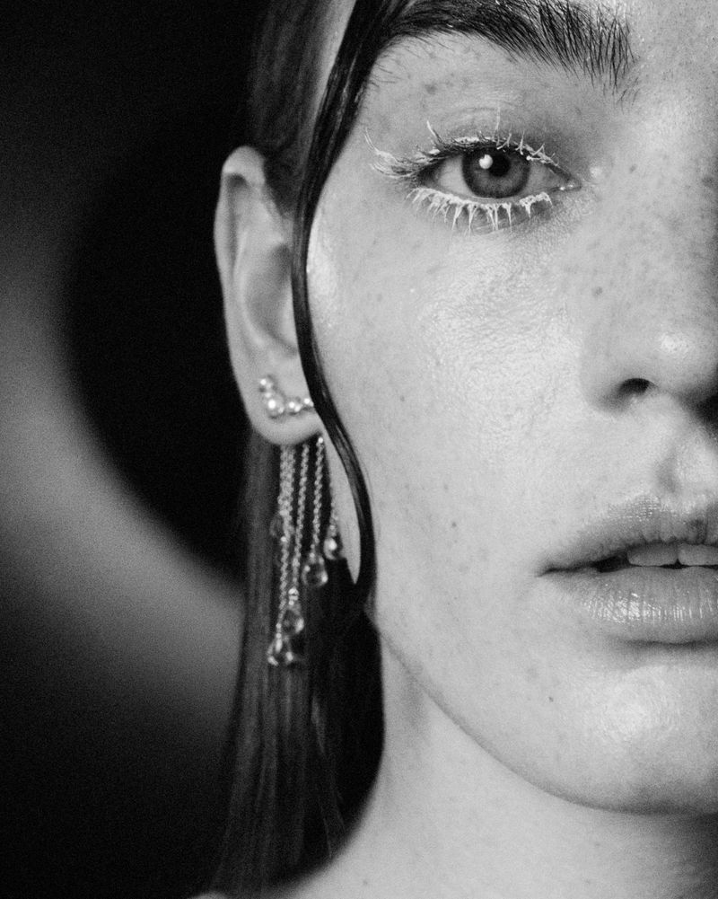 Black and white portrait of an adult woman's face, featuring her eye with white-painted eyelashes, earring, and hair strands.