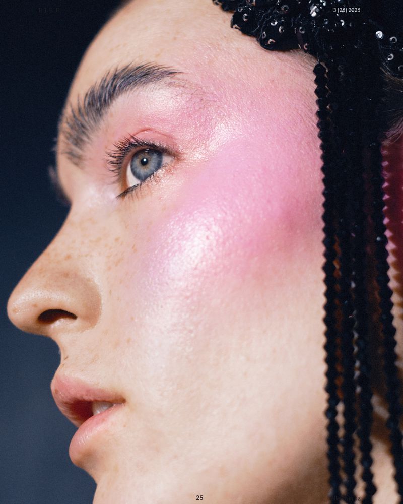 Close-up portrait of a woman with dramatic pink blush, black beaded hairpiece, freckles, and blue eyes, set against a dark background.