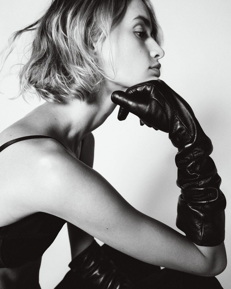 Monochrome portrait of a female model wearing long black leather gloves and resting her chin on her hand against a white backdrop.