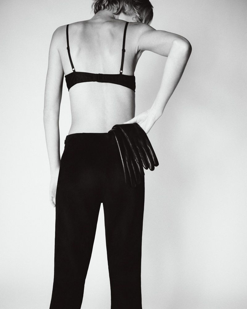 Model seen from behind wearing black trousers and holding leather gloves against a white background