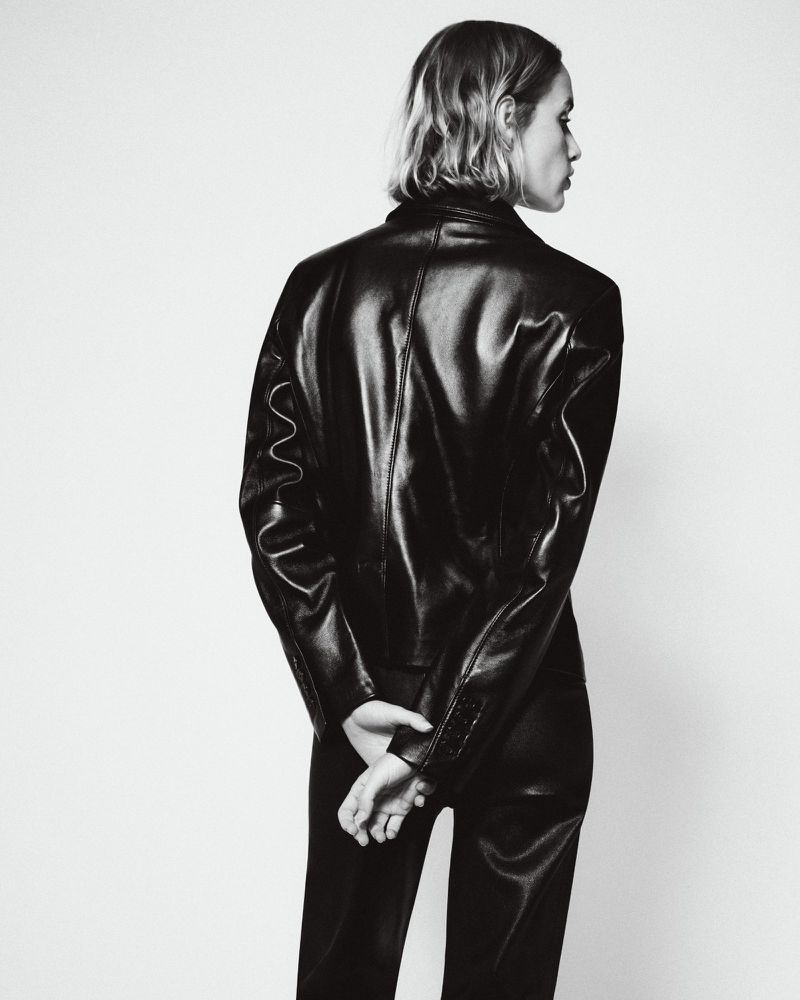 Model seen from behind wearing a black leather jacket and trousers against a white seamless backdrop.