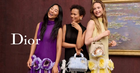 Three female models posing in colourful designer dresses holding luxury handbags against a dark textured wall with a framed painting.