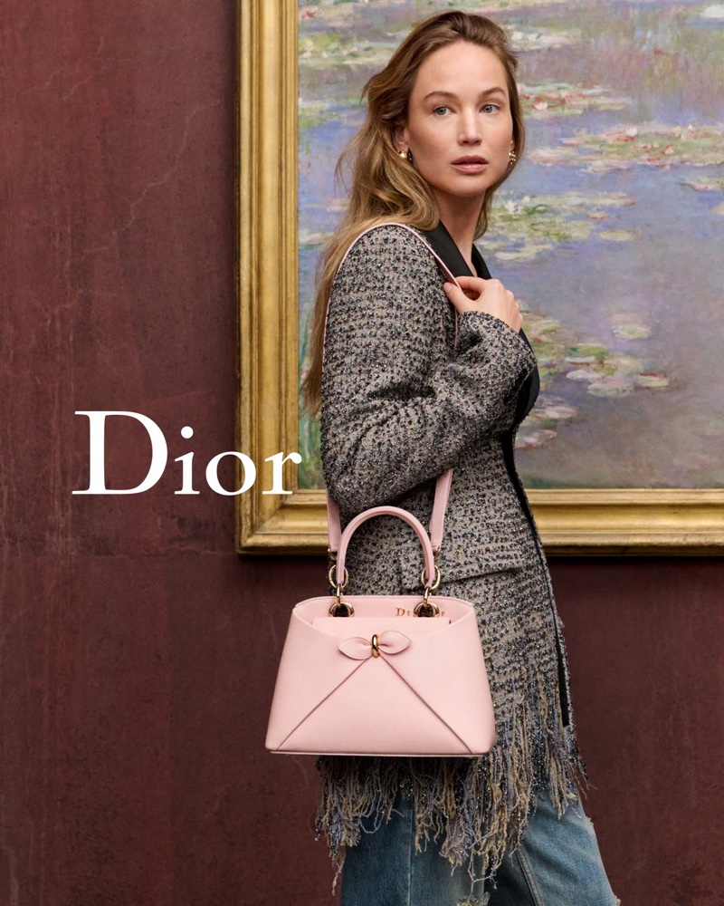 Jennifer Lawrence in a tweed jacket holding a pink Dior handbag before a framed landscape painting