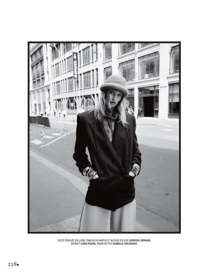Black and white editorial photo of a female model wearing a structured blazer, trousers, and a beanie on a city street.