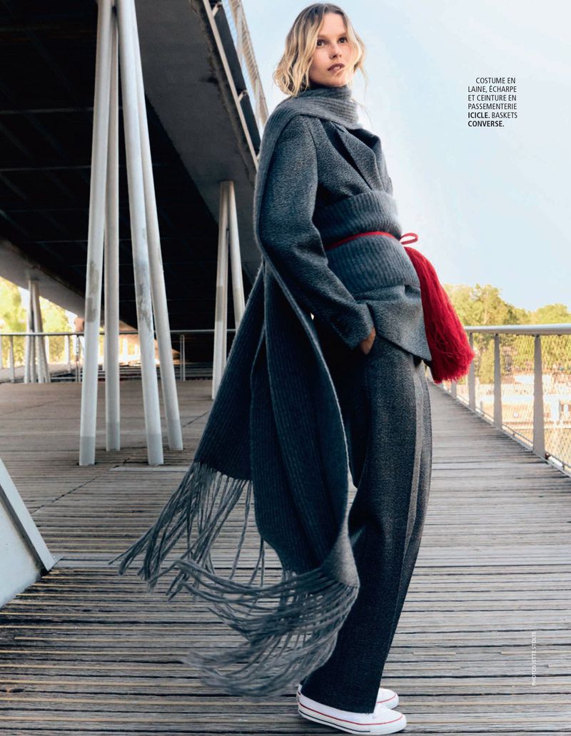 Model wearing a grey wool suit, matching scarf with fringe, and white sneakers on a wooden boardwalk beneath a concrete structure