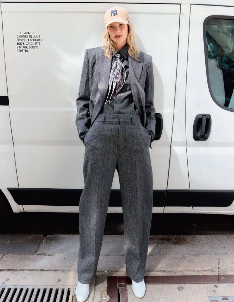 Model in grey wool suit wearing a baseball cap and patterned scarf standing against a white van