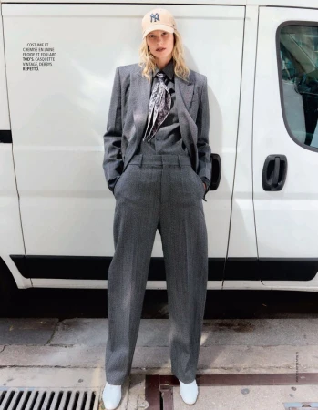 Model in grey wool suit wearing a baseball cap and patterned scarf standing against a white van