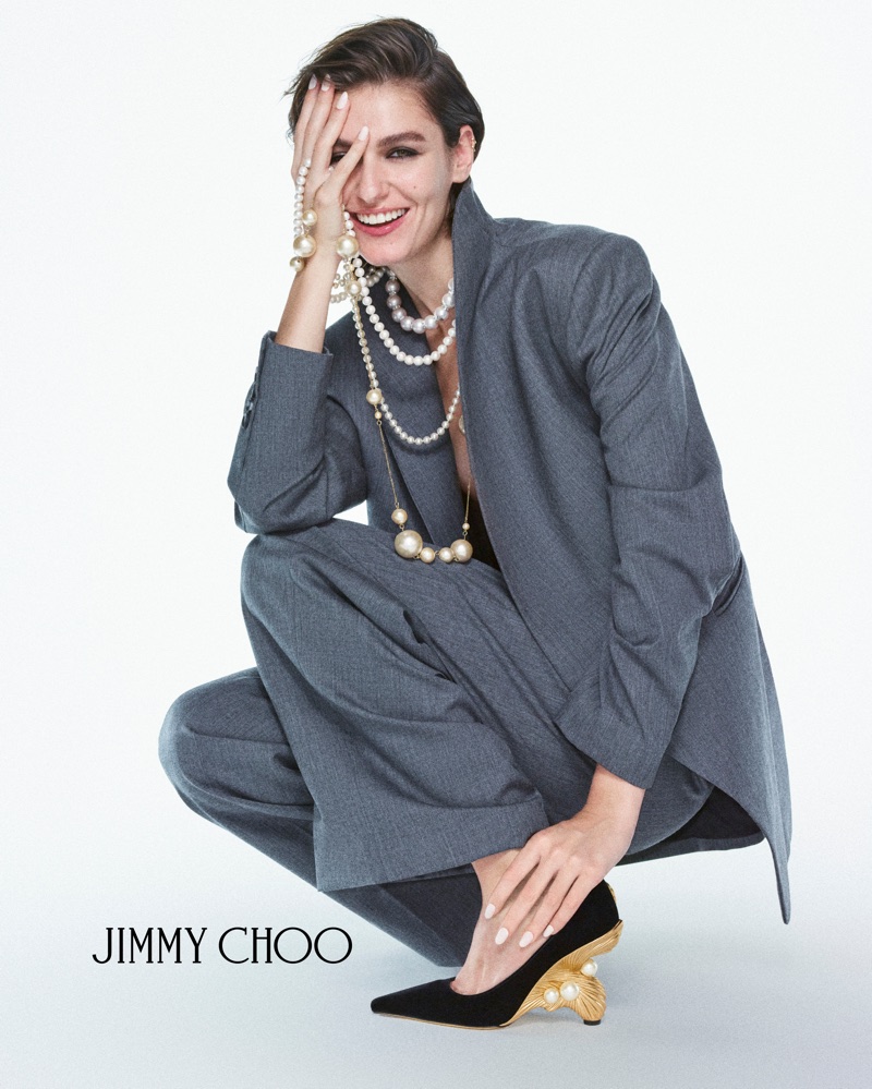 Smiling woman in grey suit crouching, wearing pearl necklaces and black heels with embellished gold heels against white seamless background