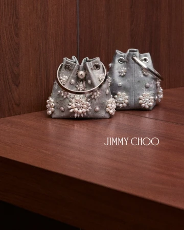 Two embellished silver bucket bags with pearl decorations resting on a dark wooden surface with Jimmy Choo branding.