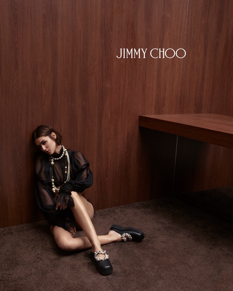 Woman seated on a brown carpet against a dark wood wall wearing a black sheer blouse, pearl necklaces, and embellished black mules.