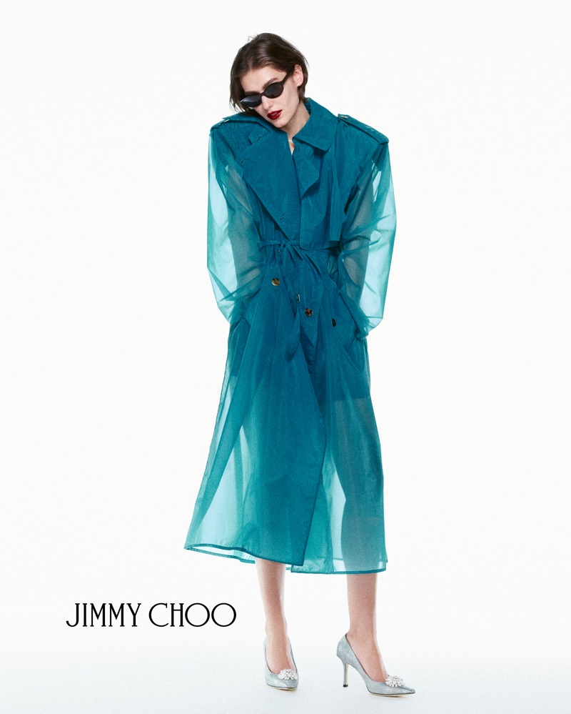 Model in dark sunglasses wearing a translucent teal coat and silver heels against a bright white background