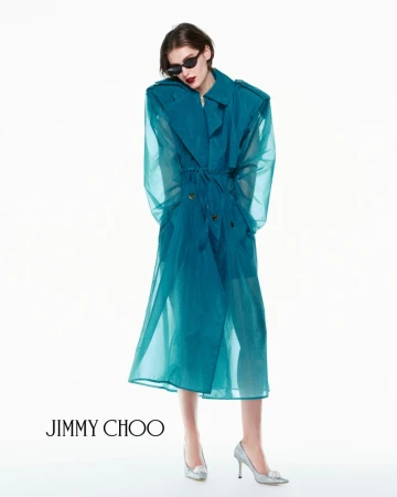 Model in dark sunglasses wearing a translucent teal coat and silver heels against a bright white background