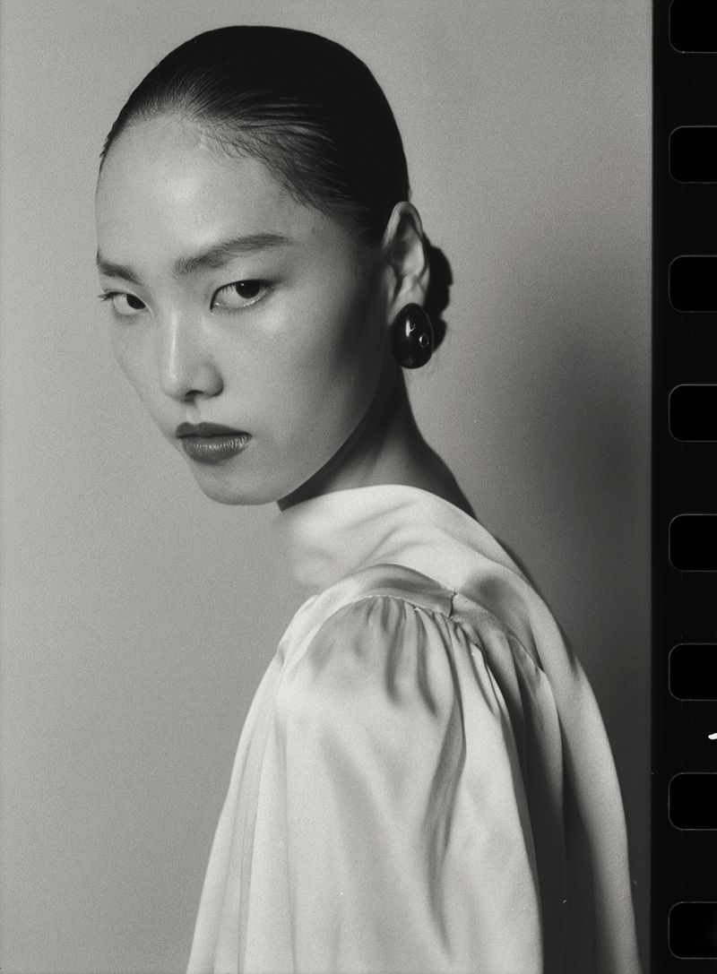 Black and white portrait of a model with hair pulled back wearing a white top and dark round earring against a neutral background.