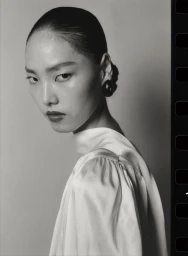 Black and white portrait of a model with hair pulled back wearing a white top and dark round earring against a neutral background.