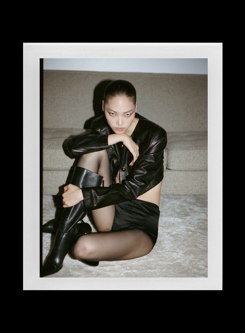 Model wearing a black leather jacket and tall boots seated on the floor against a couch