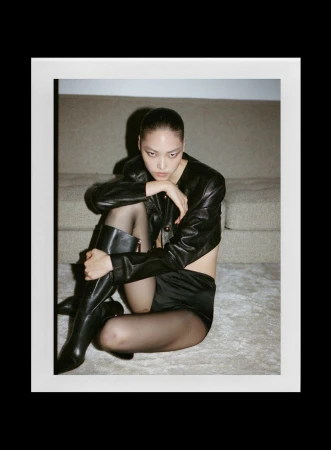 Model wearing a black leather jacket and tall boots seated on the floor against a couch
