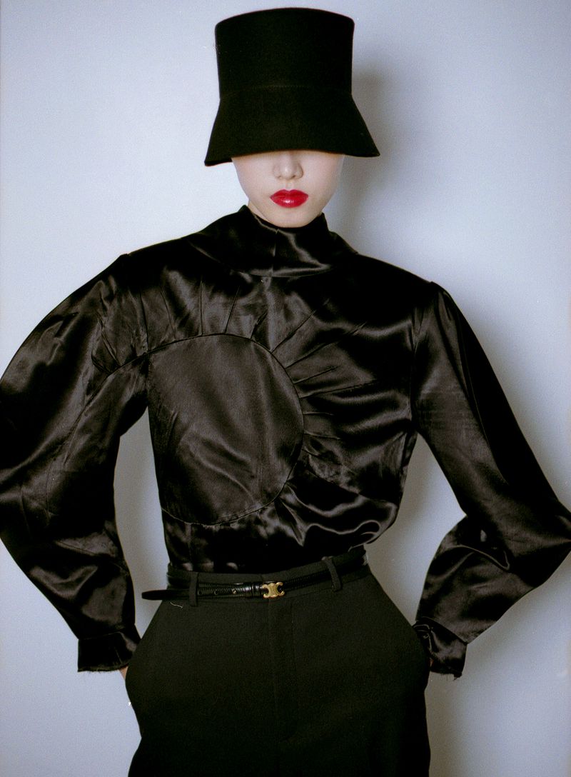 Model wearing black hat obscuring eyes, black silk top, and dark pants against white seamless background