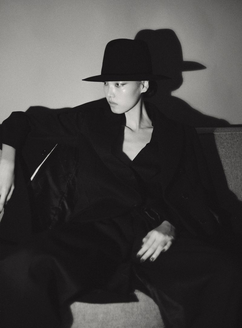 Model in black hat and dark coat seated against a wall casting a stark shadow, captured in monochrome.