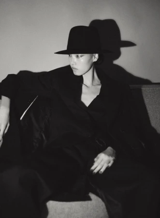 Model in black hat and dark coat seated against a wall casting a stark shadow, captured in monochrome.