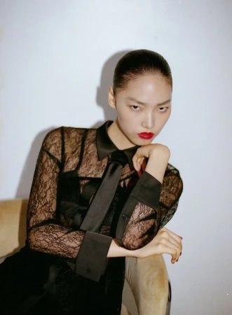 Model in black lace shirt with tie and red lipstick posing against a plain wall for Rouge Fashionbook