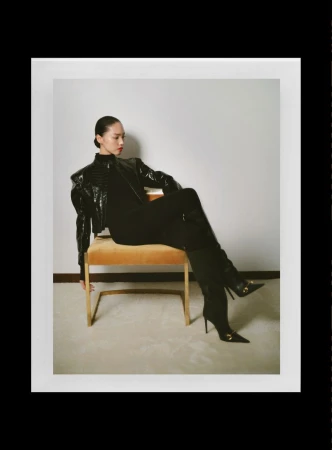 Model seated on a yellow chair wearing a black patent leather jacket and boots against a plain white wall.