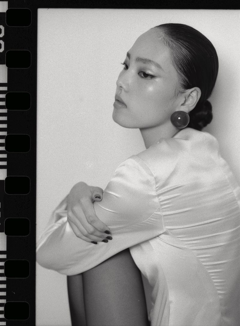 Monochrome film portrait of a model with slicked-back hair wearing a satin top and large round earring against a neutral backdrop.