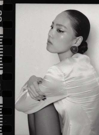 Monochrome film portrait of a model with slicked-back hair wearing a satin top and large round earring against a neutral backdrop.