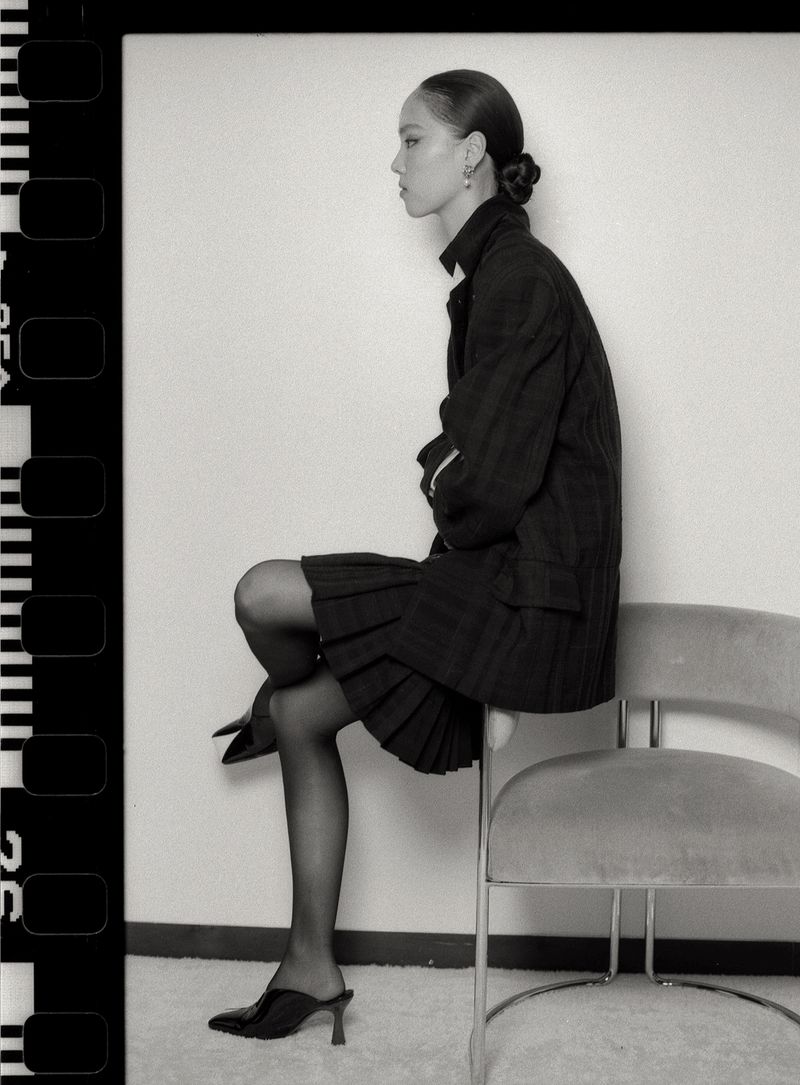 Black and white profile portrait of model Jiahui Zhang seated on an upholstered chair in a dark blazer and skirt