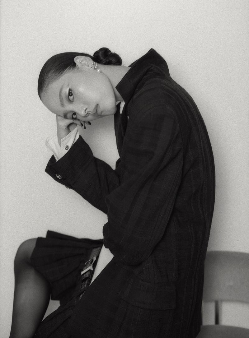 Model seated with head resting on hand, wearing a dark jacket over a pleated skirt against a plain studio background.