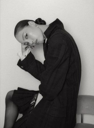 Model seated with head resting on hand, wearing a dark jacket over a pleated skirt against a plain studio background.