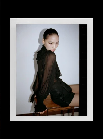 Model with slicked-back dark hair wearing a sheer black ruffled blouse sitting on a wooden stool against a white wall.