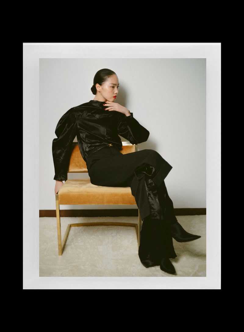 Model Jiahui Zhang seated on a golden chair wearing a black satin blouse and trousers for Rouge Fashionbook.