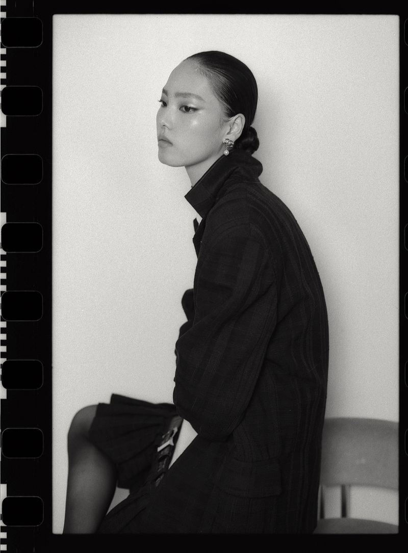 Monochrome fashion portrait of Jiahui Zhang wearing a dark jacket and skirt against a seamless white wall.