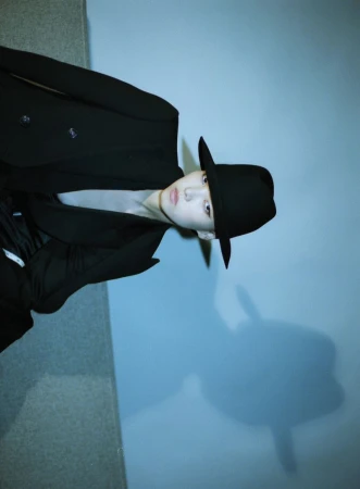 Model in black jacket and matching hat against a plain light wall with a sharp cast shadow