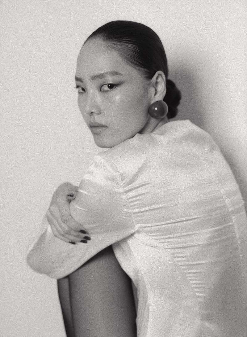 Monochrome fashion portrait of an East Asian model in a white satin top with a round dark earring looking over her shoulder.