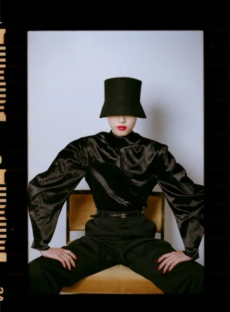 Model seated on a chair wearing a black satin blouse, trousers, belt, and a tall black hat covering the eyes.