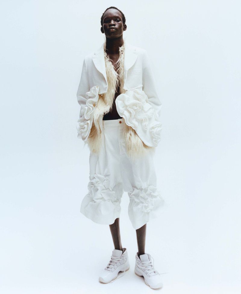 Full-length fashion portrait of a black man wearing white textured jacket with ruffles and white shorts against a white backdrop.