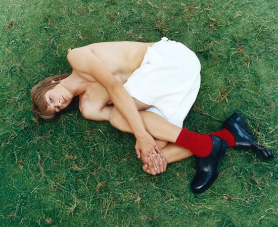 Model curled up on green grass wearing white shorts, red socks, and black shoes, photographed from above by Stef Mitchell.