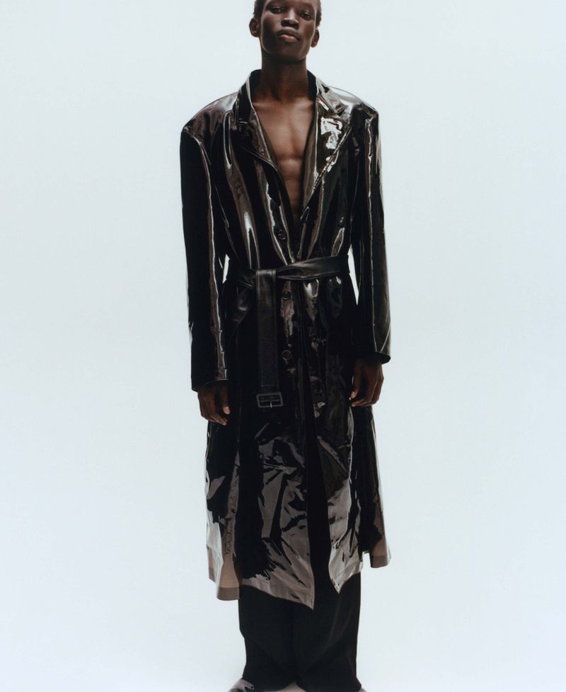 Black male model wearing a glossy dark trench coat over a bare chest against a plain light background