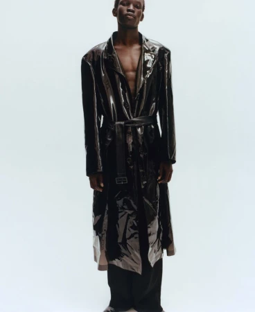 Black male model wearing a glossy dark trench coat over a bare chest against a plain light background